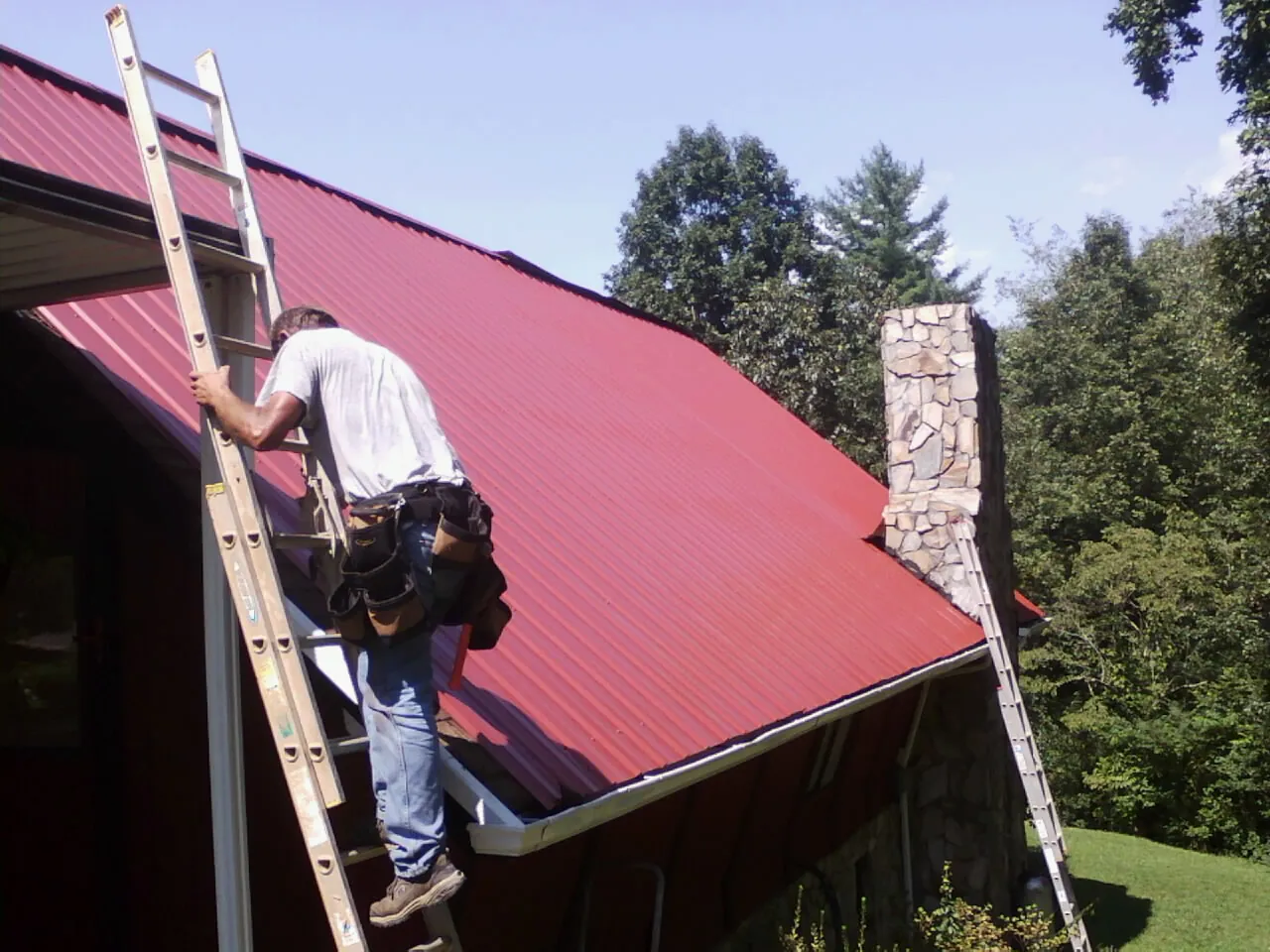 Roofer on a ladder beside a red metal roof and stone chimney — Soffit & Fascia Repair by Roofing Brothers in Buckingham